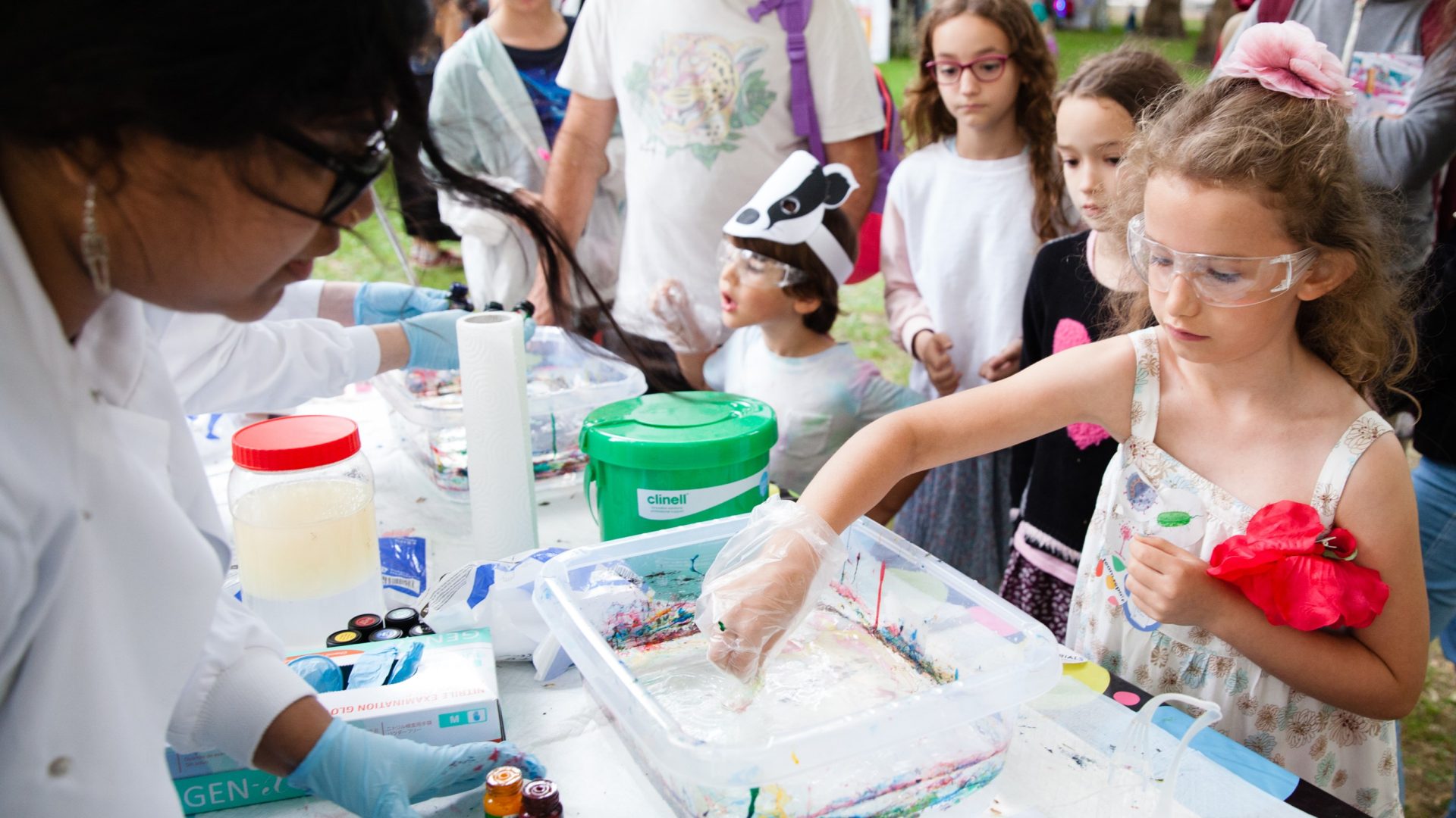 A young girl does a marbling activity at a Salters' Institute event. She is wearing a floral dress and is wearing a clear plastic glove on her right hand. She is playing in a clear plastic tub sitting on a table. The tub holds the activity for the marbling activity and some dye colours are on the table. A woman is on the other side of the table assisting the girl with the activity. The woman is wearing a lab coat and blue gloves.